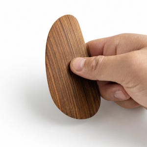 Puede incluir: Una herramienta de madera curva y semicircular con una superficie lisa y pulida que presenta un distintivo patrón de veta de madera vertical de color marrón oscuro. El objeto tiene un borde redondeado en un lado y un borde más plano y ligeramente curvo en el otro.