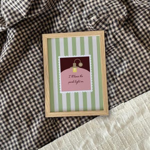 May include: A framed print with a light wood frame. The print features a green and white striped background with a square image in the center. The square image has a pink background and reads "I'll leave the porch light on."