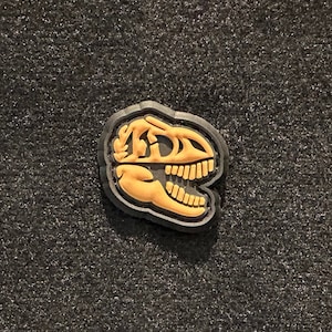May include: A black and tan dinosaur skull patch. The patch is made of fabric and has a stitched edge.