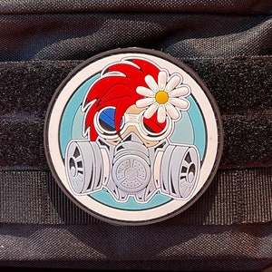 Op de afbeelding: Zwarte tactische etui met een patch met een stripfiguur met een gasmasker en een madeliefje. De patch heeft rood haar, blauwe ogen en een wit en geel madeliefje. De etui heeft een ritssluiting en klittenband.