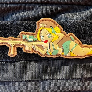 May include: A brown and yellow embroidered patch depicting a cartoon woman in a green and yellow outfit holding a rifle. The woman is wearing a green hat with a yellow insignia. The patch is attached to a black tactical vest.
