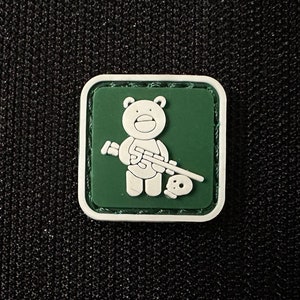Op de afbeelding: Een vierkante patch met een groene achtergrond en een witte rand. De patch toont een cartoonbeer met een geweer en een schedel. De beer is wit en het geweer en de schedel zijn ook wit.