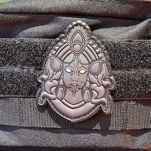 Könnte beinhalten: Eine schwarze taktische Tasche mit einem silbernen Metallabzeichen, das eine detaillierte Illustration einer Frau mit einem dritten Auge und herausgestreckter Zunge zeigt.