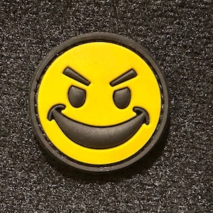 May include: A yellow smiley face patch with a black outline and a mischievous grin. The patch has a black border.