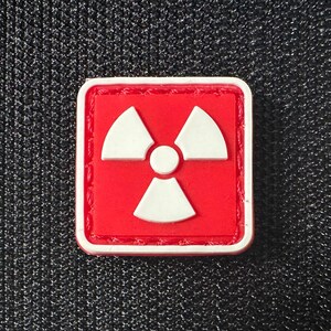 Könnte beinhalten: Ein roter quadratischer Patch mit einem weißen Strahlungssymbol. Das Symbol ist ein stilisiertes dreiblättriges Kleeblatt mit einem Kreis in der Mitte.