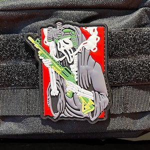 Pode incluir: Bolsa tática preta com um remendo de velcro que apresenta um esqueleto vestindo uma túnica com capuz e segurando um rifle. O remendo tem um fundo vermelho com detalhes verdes e amarelos.