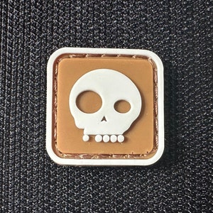 May include: A brown and white skull patch with a white outline. The patch is made of a soft, textured material and has a stitched edge.