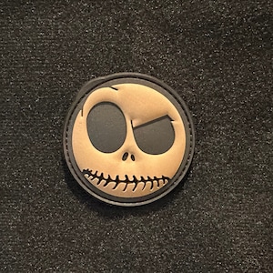 May include: A black and tan embroidered patch featuring the face of Jack Skellington from the movie "The Nightmare Before Christmas".