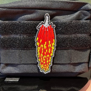 May include: A black tactical pouch with a red and yellow embroidered patch of a bundle of dynamite sticks. The patch is attached to the front of the pouch with velcro.