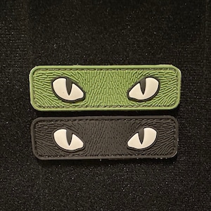 May include: Two rectangular patches with white eyes on a black and green background. The patches are made of a textured material and have a stitched edge.