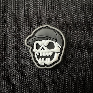 Op de afbeelding: Een zwart-witte schedel met een zwarte baseballpet. De schedel heeft een witte glimlach en witte ogen. De schedel is op een zwarte achtergrond.