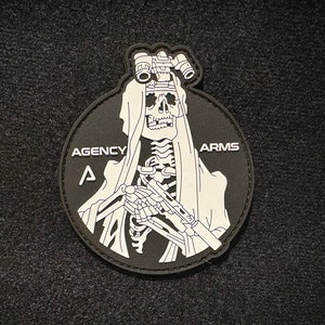 May include: A black and white patch with a skeleton wearing a hooded cloak and holding a gun. The skeleton is wearing binoculars on its head. The text "AGENCY ARMS" is printed on the patch.