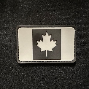 Puede incluir: Un parche de bandera canadiense en blanco y negro con una hoja de arce blanca sobre un fondo gris. El parche tiene un borde negro y está hecho de un material texturizado.