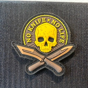 May include: A yellow and brown embroidered patch with a skull and crossbones. The patch has the text "NO KNIFE NO LIFE" around the skull and "BACKHAND" on the crossbones.