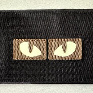 May include: Two brown patches with a white glow-in-the-dark eye design, sewn onto a black fabric background.