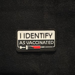May include: A black and white patch with the text "I IDENTIFY AS VACCINATED" and a stylized image of a syringe with a red fill line.