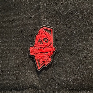 May include: Red embroidered patch with a skull wearing a hooded cloak and biting down on a bomb. The patch is on a black background.