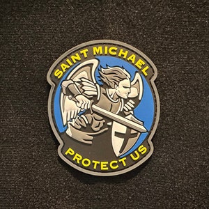 Puede incluir: Un parche con una ilustración de San Miguel, con una espada y un escudo, sobre un fondo azul y gris. El texto "SAINT MICHAEL PROTECT US" está en amarillo y blanco.