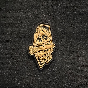 May include: A gold-colored patch featuring a skull wearing a hood and holding a bomb. The patch has a black border and is set against a dark background. The skull has detailed features.