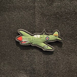 Pode incluir: Um patch verde em forma de avião com um nariz vermelho e um design de boca de tubarão. O patch tem uma borda preta e o número "113521" na cauda. O avião tem um emblema de estrela na asa.