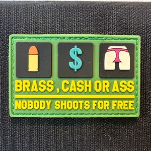 May include: A rectangular patch with a green border and black background. The patch features three icons: a bullet, a dollar sign, and a cartoon rear end. Text reads "BRASS, CASH OR ASS" and "NOBODY SHOOTS FOR FREE."
