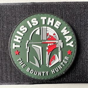 May include: A green, white and red circular patch with the text "This is the way The Bounty Hunter" and a stylised image of a Mandalorian helmet.