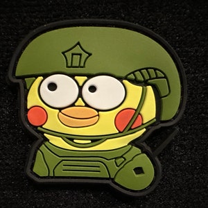Peut inclure: Écusson de style dessin animé représentant un personnage jaune portant un casque et un uniforme militaires verts. Le personnage a de grands yeux blancs, des joues oranges et une expression souriante. Le patch est bordé de noir.