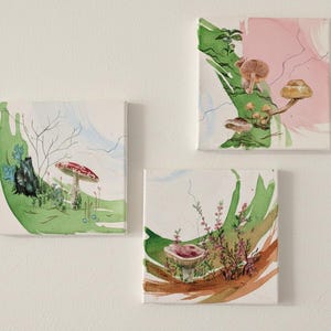 May include: Three square canvas art pieces featuring watercolor paintings of mushrooms and foliage. The paintings have a whimsical style with a palette of green, pink, and brown. The art is mounted on a white wall.