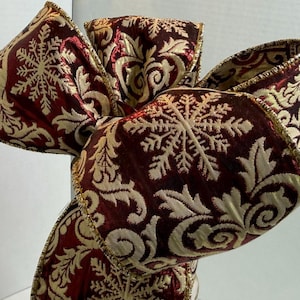May include: A decorative bow made from wired ribbon featuring a deep burgundy red base with a metallic gold woven pattern of intricate snowflakes and swirling acanthus leaves. The edges of the ribbon are finished with a thin gold metallic trim.