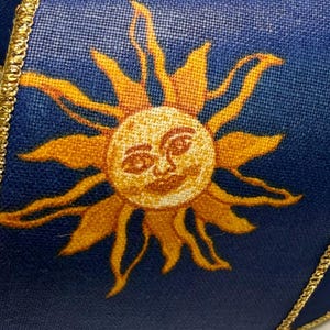 May include: A navy blue wired ribbon featuring a repeating pattern of golden yellow suns with faces and small white stars. The fabric ribbon has metallic gold finished edges and is wound around a white spool, showing a textured weave surface.