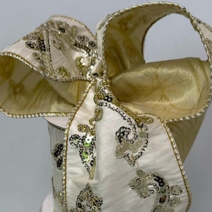 May include: A decorative ribbon featuring a cream-colored fabric base with intricate gold sequin embroidery in a swirling pattern. The ribbon has a wired edge finished in gold thread, allowing it to be shaped into a large, voluminous bow.