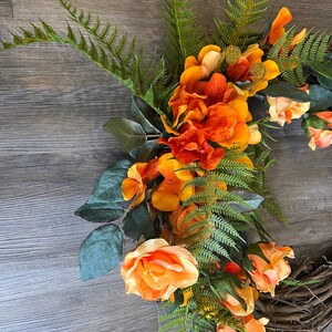 May include: A wreath made of grapevine with orange and yellow flowers and green ferns.