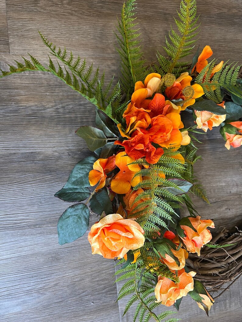 May include: A wreath made of grapevine with orange and yellow flowers and green ferns.
