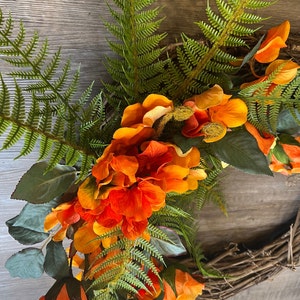 May include: A wreath made of grapevine branches decorated with orange and yellow silk flowers and green fern leaves.