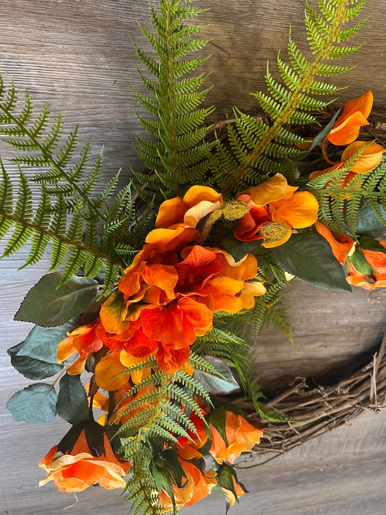 May include: A wreath made of grapevine branches decorated with orange and yellow silk flowers and green fern leaves.
