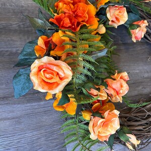 May include: A wreath made of grapevine with orange and peach colored roses and orange flowers with green fern leaves.