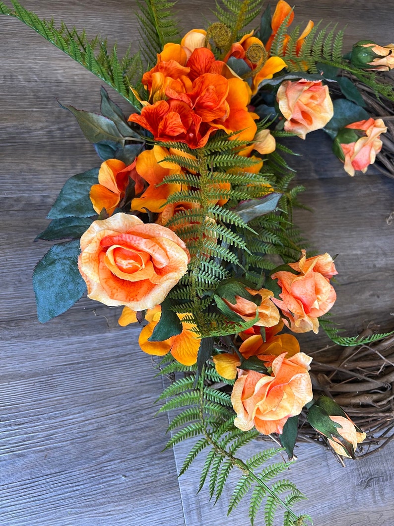 May include: A wreath made of grapevine with orange and peach colored roses and orange flowers with green fern leaves.