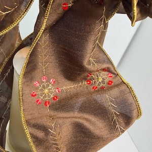 May include: A brown fabric ribbon featuring gold-colored stitched branch patterns and small, circular red sequins arranged in floral-like clusters. The edges of the ribbon are finished with a metallic gold-colored thread trim.