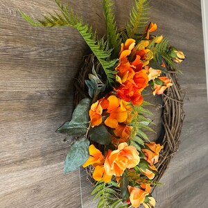 May include: A wreath made of grapevine with orange and peach colored flowers and green ferns.