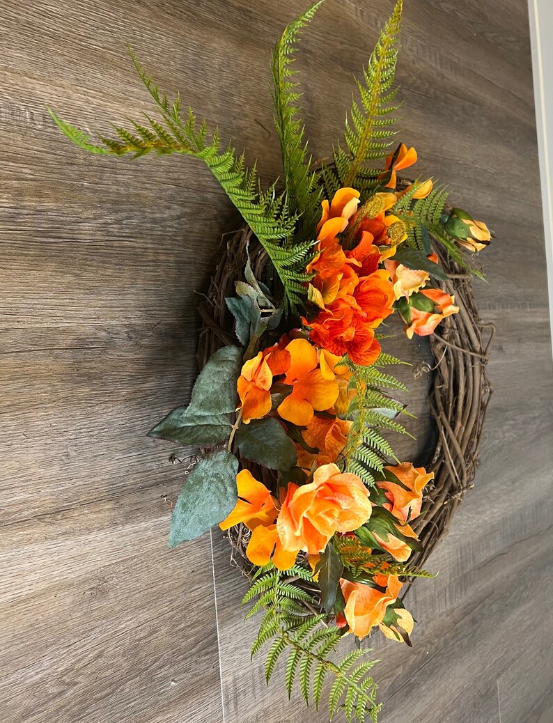 May include: A wreath made of grapevine with orange and peach colored flowers and green ferns.