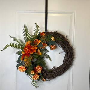 May include: A brown grapevine wreath decorated with orange and yellow flowers and green ferns.
