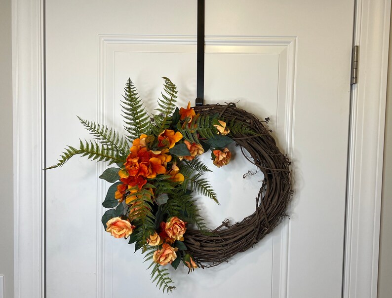 May include: A brown grapevine wreath decorated with orange and yellow flowers and green ferns.