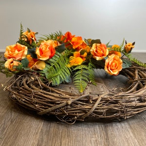 May include: A brown grapevine wreath decorated with orange and yellow artificial roses and green fern leaves.
