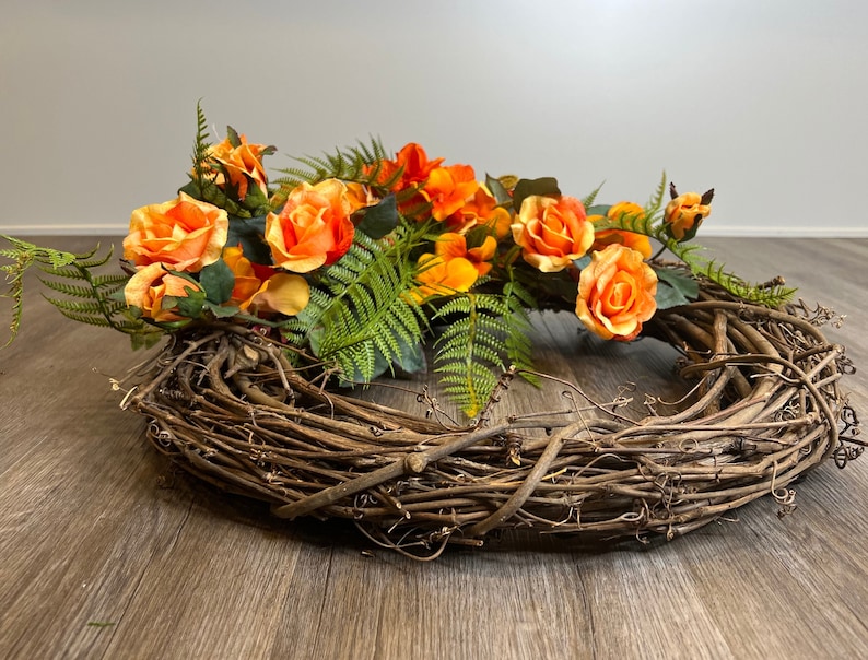 May include: A brown grapevine wreath decorated with orange and yellow artificial roses and green fern leaves.