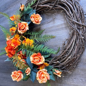 May include: A grapevine wreath decorated with orange and yellow roses and green ferns.