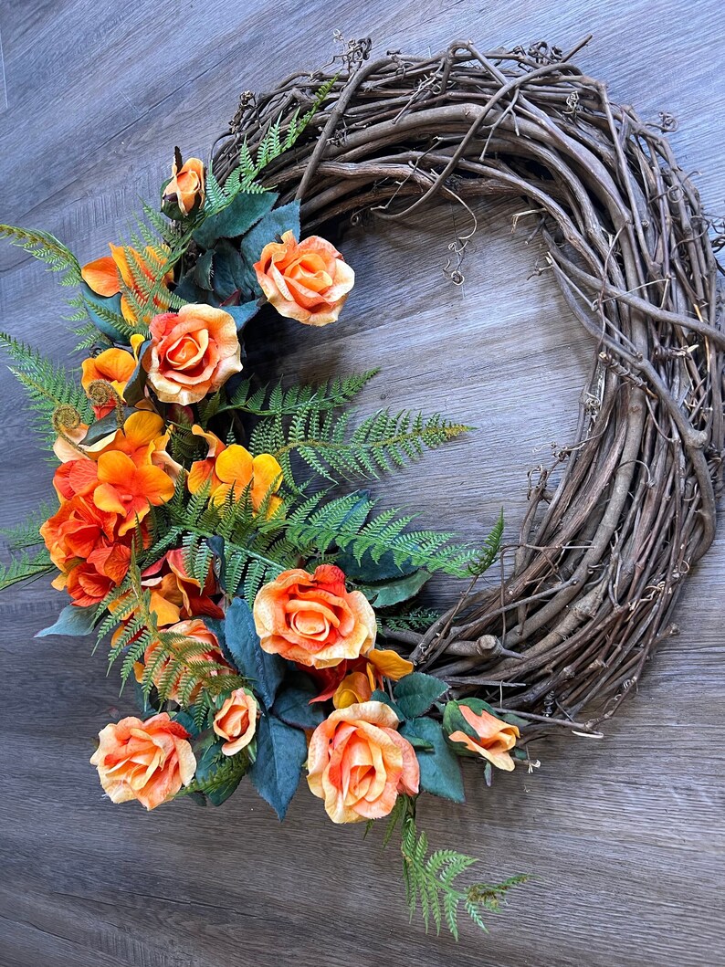 May include: A grapevine wreath decorated with orange and yellow roses and green ferns.