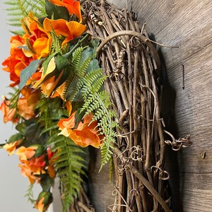 May include: A wreath made of brown grapevine branches with orange and yellow silk flowers and green fern leaves.