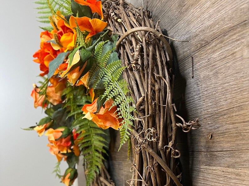 May include: A wreath made of brown grapevine branches with orange and yellow silk flowers and green fern leaves.