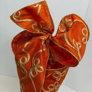 May include: A decorative bow crafted from vibrant orange fabric featuring a metallic gold embroidered swirl pattern. The edges of the bow are finished with a thin gold wire trim, creating a structured shape with multiple loops and flowing ribbon ends.