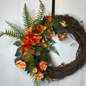 May include: A brown grapevine wreath decorated with orange and yellow flowers and green ferns.